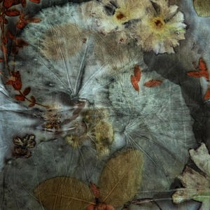 May include: A textile piece featuring botanical eco-printing with visible leaf and flower imprints. The fabric displays a muted color palette of grey, brown, and rust orange, showcasing intricate vein patterns from various foliage and floral shapes.