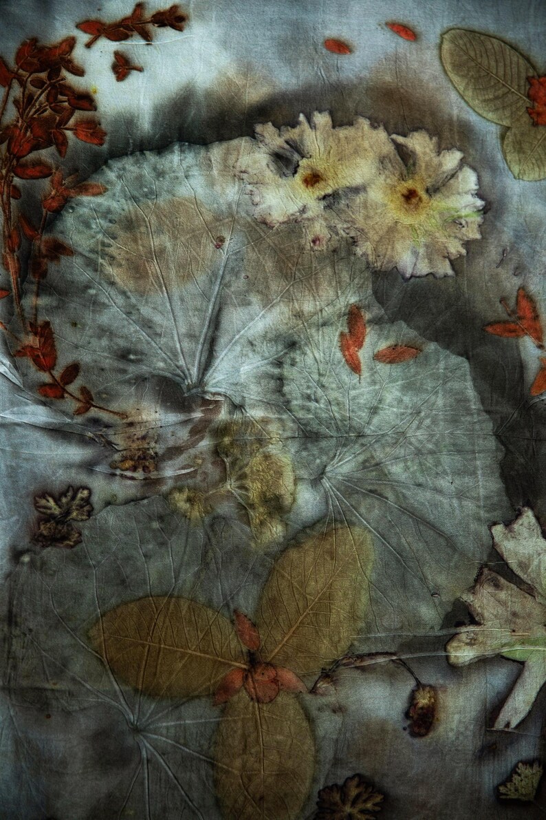 May include: A textile piece featuring botanical eco-printing with visible leaf and flower imprints. The fabric displays a muted color palette of grey, brown, and rust orange, showcasing intricate vein patterns from various foliage and floral shapes.