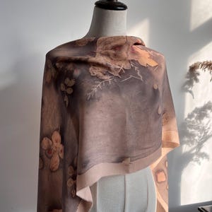 May include: A rectangular scarf featuring a botanical print with muted brown, grey, and beige tones. The fabric displays detailed imprints of various leaves and flowers scattered across the surface, with a light beige border along the edges.