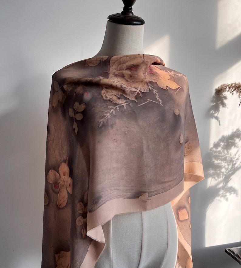 May include: A rectangular scarf featuring a botanical print with muted brown, grey, and beige tones. The fabric displays detailed imprints of various leaves and flowers scattered across the surface, with a light beige border along the edges.