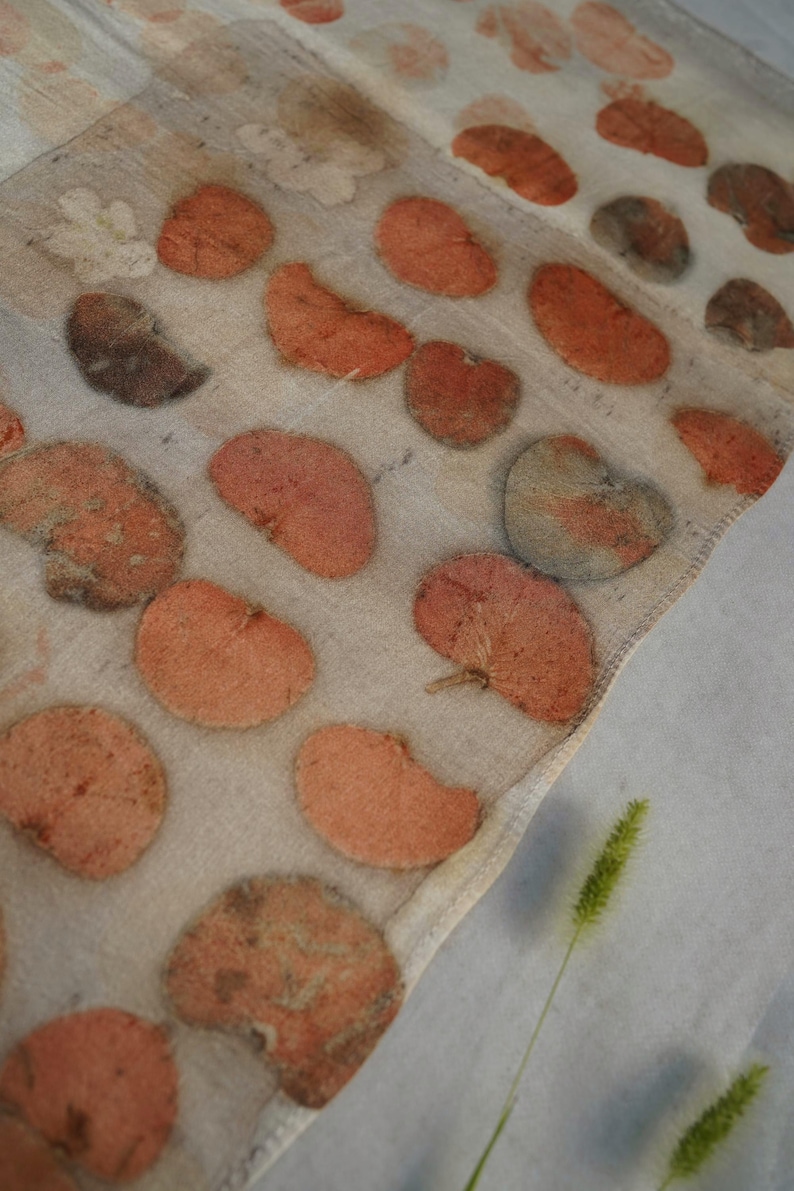 May include: A lightweight, semi-sheer off-white scarf featuring a botanical print of scattered, rounded leaves in shades of terracotta, muted orange, and soft brown. The fabric displays a natural, organic texture with visible leaf vein patterns.