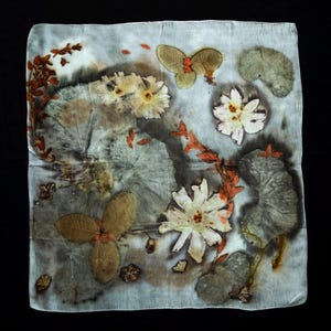 May include: A square silk scarf featuring a botanical print with pressed flowers and leaves in shades of white, muted green, and burnt orange. The fabric displays a natural, organic pattern of various plant shapes, including large rounded leaves and delicate floral blooms, set against a pale, light gray background.