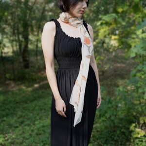 以下が含まれることがあります： 黒いノースリーブのドレスと、オレンジ色の花柄のクリーム色のスカーフを身につけた女性。 ドレスはフィットした胴体と流れるようなスカートです。 背景はぼやけた森です。