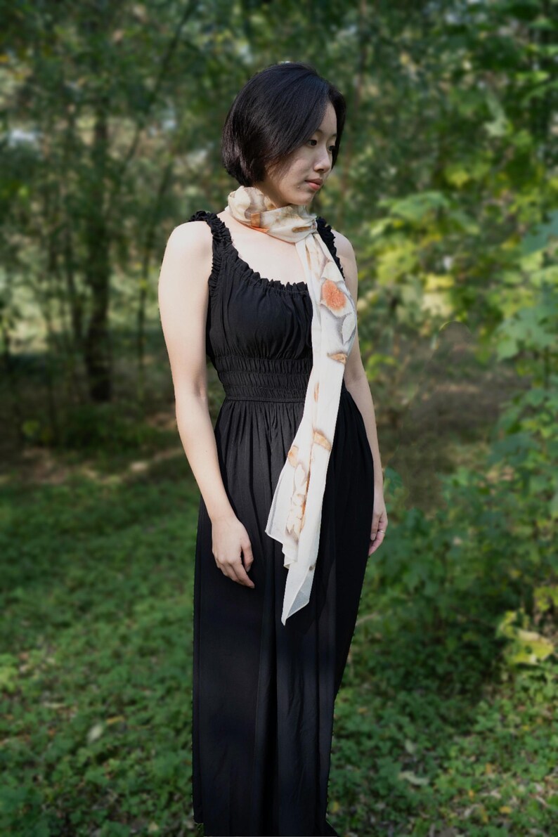 以下が含まれることがあります： 黒いノースリーブのドレスと、オレンジ色の花柄のクリーム色のスカーフを身につけた女性。 ドレスはフィットした胴体と流れるようなスカートです。 背景はぼやけた森です。