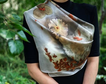 May include: A rectangular silk scarf featuring a botanical print with muted gray and white tones, accented by pressed flower and leaf patterns in shades of rust, orange, and pale yellow, draped over a solid black short-sleeved shirt.