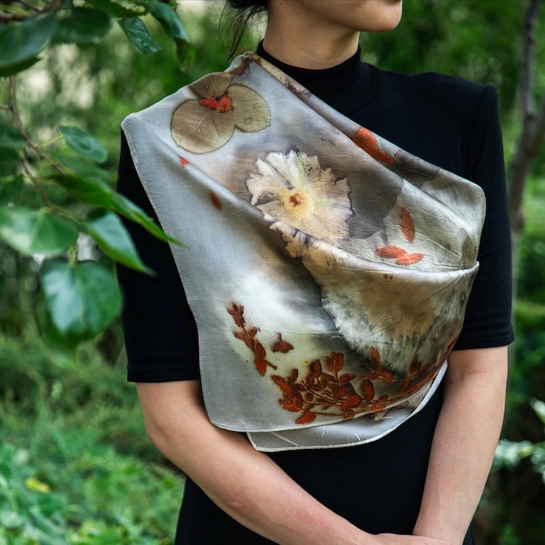 May include: A rectangular silk scarf featuring a botanical print with muted gray and white tones, accented by pressed flower and leaf patterns in shades of rust, orange, and pale yellow, draped over a solid black short-sleeved shirt.