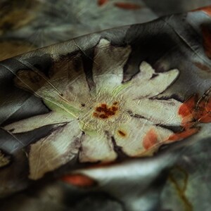 May include: A piece of fabric featuring a botanical print with large, pale, multi-petaled flowers and dark, moody foliage. The design includes muted green, brown, and orange tones, with a soft, draped texture that creates deep shadows and highlights.