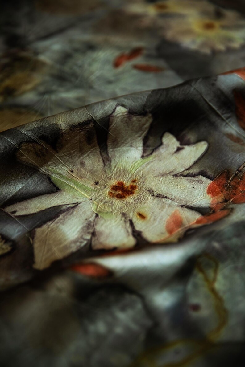 May include: A piece of fabric featuring a botanical print with large, pale, multi-petaled flowers and dark, moody foliage. The design includes muted green, brown, and orange tones, with a soft, draped texture that creates deep shadows and highlights.