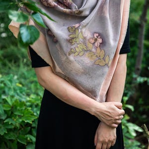 May include: A large, rectangular scarf featuring a muted lavender and soft peach color palette with botanical imprints of leaves and flowers in shades of green and brown. The fabric has raw, unfinished edges and a smooth, draped texture.