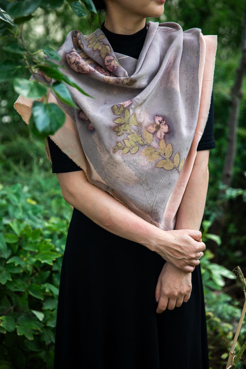 May include: A large, rectangular scarf featuring a muted lavender and soft peach color palette with botanical imprints of leaves and flowers in shades of green and brown. The fabric has raw, unfinished edges and a smooth, draped texture.