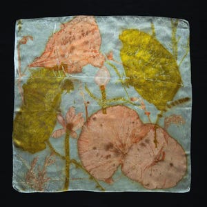 May include: A square silk scarf featuring a botanical print with large, rounded peach-colored leaves and vibrant yellow foliage against a pale, translucent grey background. The design includes delicate stems and small floral elements.