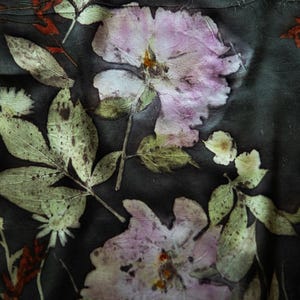 May include: A dark charcoal fabric featuring a botanical print of pressed pink flowers and green leaves. The design displays detailed textures of petals and foliage scattered across the surface, creating a natural, organic pattern on the textile.