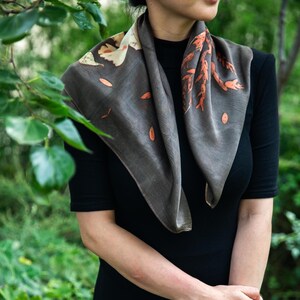 May include: A square scarf in a dark charcoal gray hue featuring botanical prints of leaves and branches in shades of cream and burnt orange. The scarf is draped around the neck, showcasing a smooth texture and a natural, organic leaf pattern.