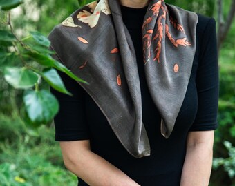Puede incluir: Un pañuelo cuadrado de color gris carbón oscuro con estampados botánicos de hojas y ramas en tonos crema y naranja quemado. El pañuelo está drapeado alrededor del cuello, mostrando una textura suave y un patrón de hojas natural.