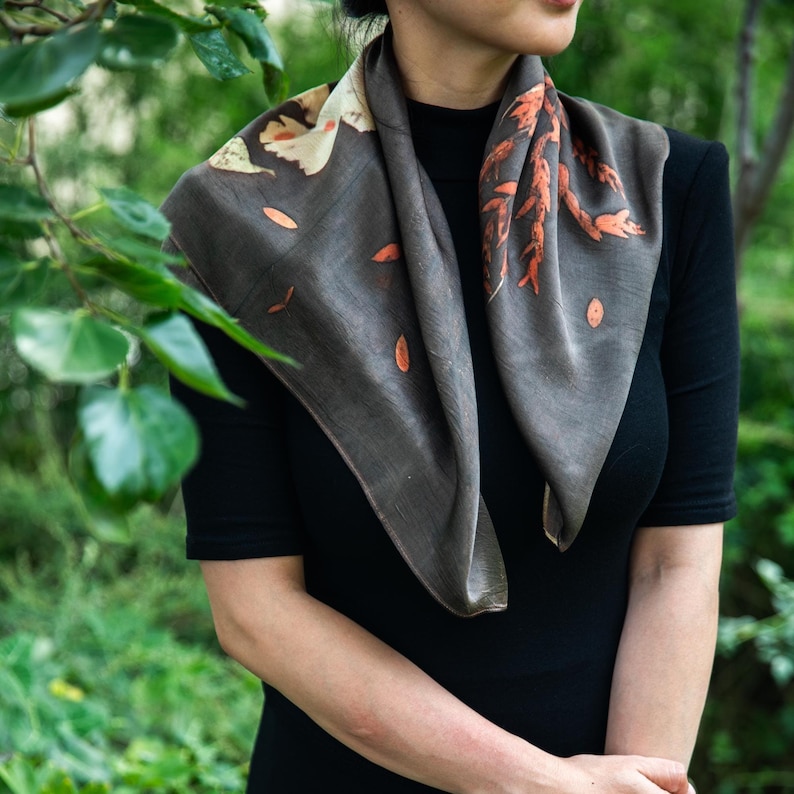 May include: A square scarf in a dark charcoal gray hue featuring botanical prints of leaves and branches in shades of cream and burnt orange. The scarf is draped around the neck, showcasing a smooth texture and a natural, organic leaf pattern.