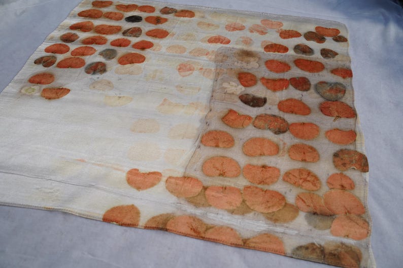 May include: A rectangular off-white textile featuring a repeating pattern of pressed orange and brown leaf prints arranged in rows. The fabric has a semi-sheer appearance with visible botanical imprints that vary in color intensity and shape.