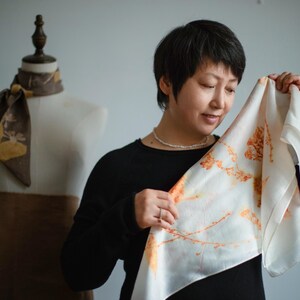 May include: A square white silk scarf featuring a botanical print of orange and yellow plant stems and leaves. The scarf is held up to display the delicate floral pattern, which contrasts with the smooth, light-reflecting surface of the fabric.