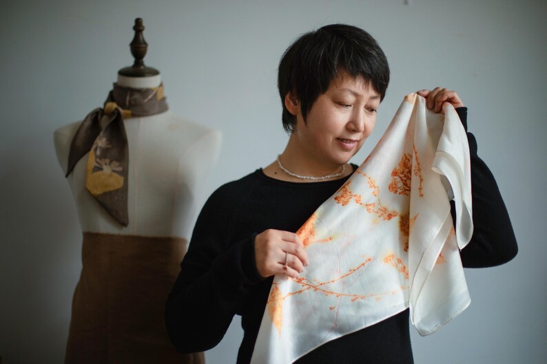 May include: A square white silk scarf featuring a botanical print of orange and yellow plant stems and leaves. The scarf is held up to display the delicate floral pattern, which contrasts with the smooth, light-reflecting surface of the fabric.