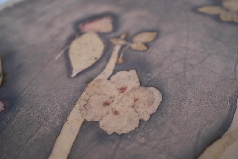 May include: A piece of fabric featuring a botanical print with light beige flower and leaf silhouettes against a dark, muted grey background. The surface displays a visible crackled texture throughout, with subtle variations in colour depth.