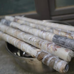 May include: A collection of long, cylindrical items wrapped in clear plastic film, resting inside a metallic bowl. The items appear to have a textured, brownish-grey surface visible through the transparent wrapping, arranged in a parallel stack.
