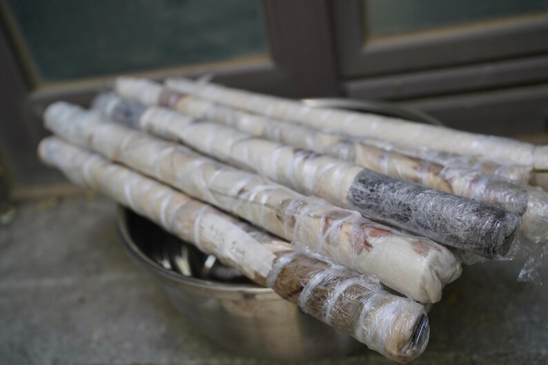 May include: A collection of long, cylindrical items wrapped in clear plastic film, resting inside a metallic bowl. The items appear to have a textured, brownish-grey surface visible through the transparent wrapping, arranged in a parallel stack.
