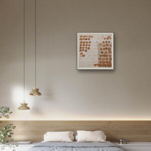K&ouml;nnte beinhalten: Ein quadratisches gerahmtes Wandbild mit einer gitterartigen Anordnung kleiner, kreisf&ouml;rmiger, rostfarbener und brauner strukturierter Formen auf hellem, cremewei&szlig;em Hintergrund, pr&auml;sentiert in einem schlichten, d&uuml;nnen, dunklen Rahmen.