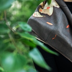 May include: A dark charcoal gray scarf featuring a botanical print with scattered orange and cream-colored leaf motifs. The fabric has a smooth, matte texture and is draped over the shoulders, displaying a natural, organic pattern of foliage.