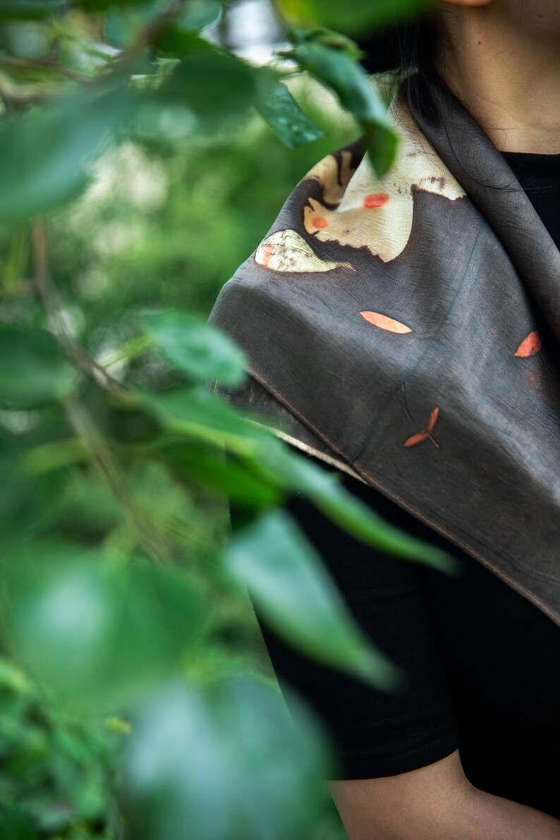 May include: A dark charcoal gray scarf featuring a botanical print with scattered orange and cream-colored leaf motifs. The fabric has a smooth, matte texture and is draped over the shoulders, displaying a natural, organic pattern of foliage.