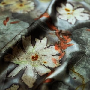 May include: A grey silk scarf featuring botanical prints of white flowers and orange leaves. The fabric has a soft, draped texture with visible plant imprints, including large white petals and delicate foliage patterns scattered across the surface.