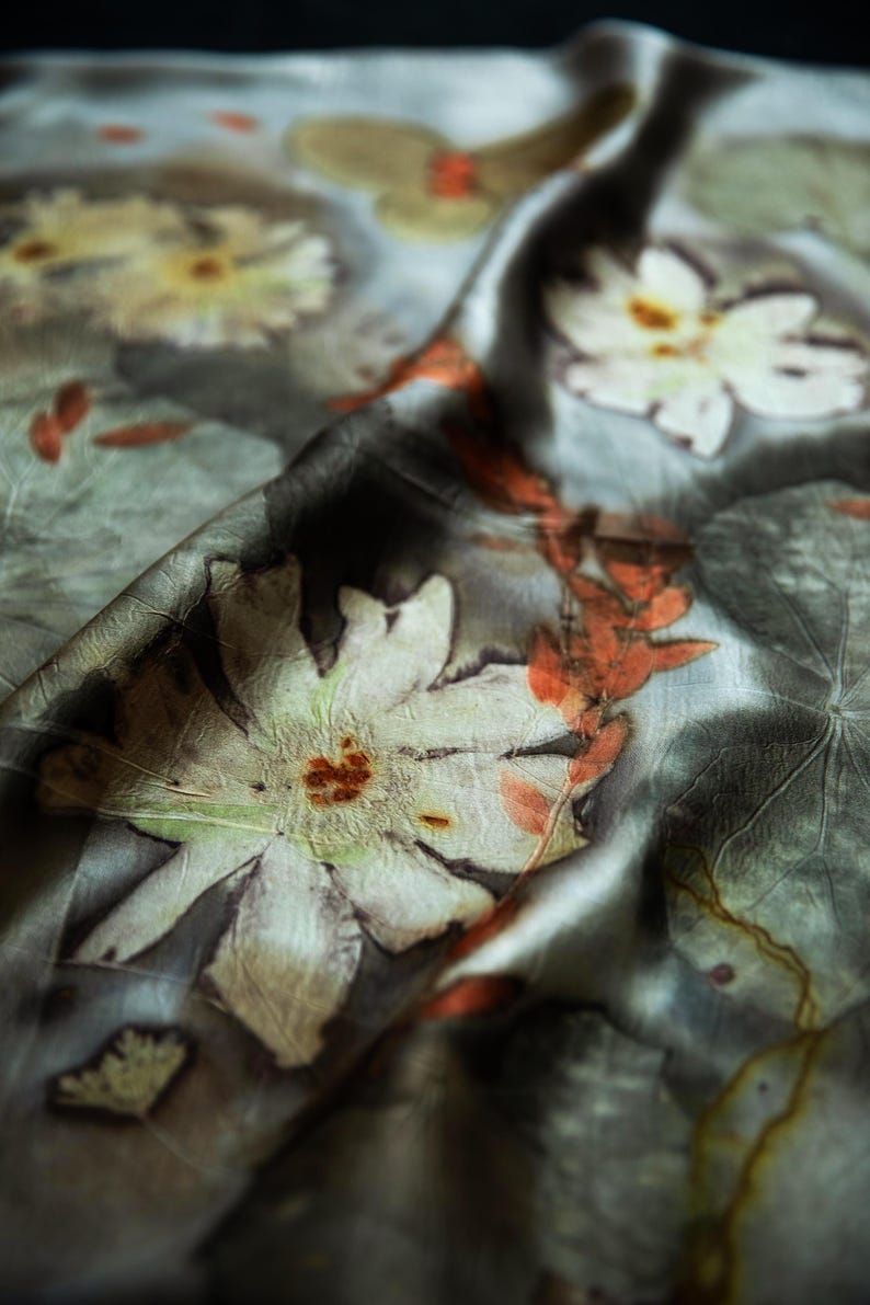 May include: A grey silk scarf featuring botanical prints of white flowers and orange leaves. The fabric has a soft, draped texture with visible plant imprints, including large white petals and delicate foliage patterns scattered across the surface.