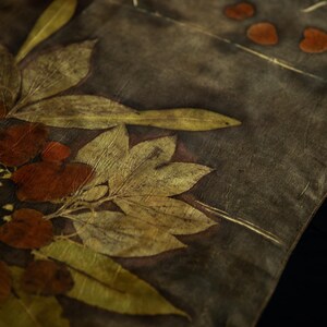 May include: A textile piece featuring a dark gray background with botanical imprints of elongated yellow-green leaves and rounded reddish-brown foliage. The fabric displays a natural, organic pattern created through leaf contact printing techniques.