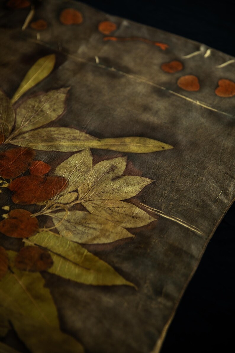May include: A textile piece featuring a dark gray background with botanical imprints of elongated yellow-green leaves and rounded reddish-brown foliage. The fabric displays a natural, organic pattern created through leaf contact printing techniques.