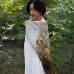 May include: A rectangular scarf featuring a botanical print with yellow flower and dark brown foliage patterns on a light gray background, draped over a plain white garment. The fabric displays a gradient effect transitioning from light gray to dark brown.