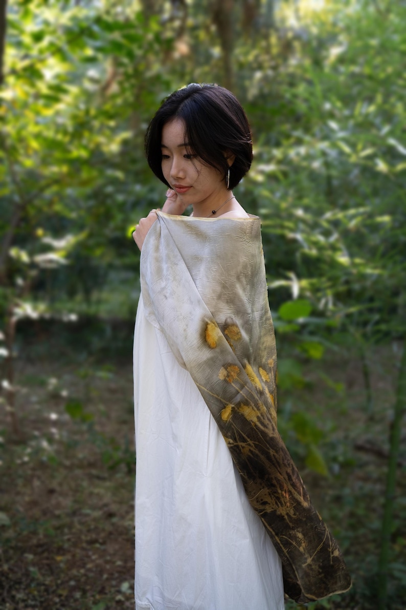 May include: A rectangular scarf featuring a botanical print with yellow flower and dark brown foliage patterns on a light gray background, draped over a plain white garment. The fabric displays a gradient effect transitioning from light gray to dark brown.