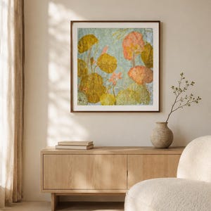 May include: A square wall art piece in a dark brown frame featuring a botanical print with pressed yellow, orange, and green leaves and flowers against a light blue background, displayed above a light wood sideboard with two books and a vase.