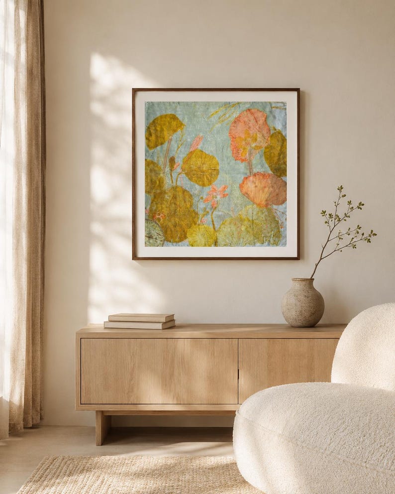 May include: A square wall art piece in a dark brown frame featuring a botanical print with pressed yellow, orange, and green leaves and flowers against a light blue background, displayed above a light wood sideboard with two books and a vase.