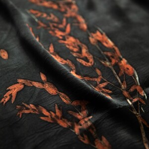 May include: A dark charcoal gray fabric featuring a botanical print of elongated, rust-colored leaves and stems. The material has a soft, slightly wrinkled texture with folds that create depth and shadows across the surface of the textile.