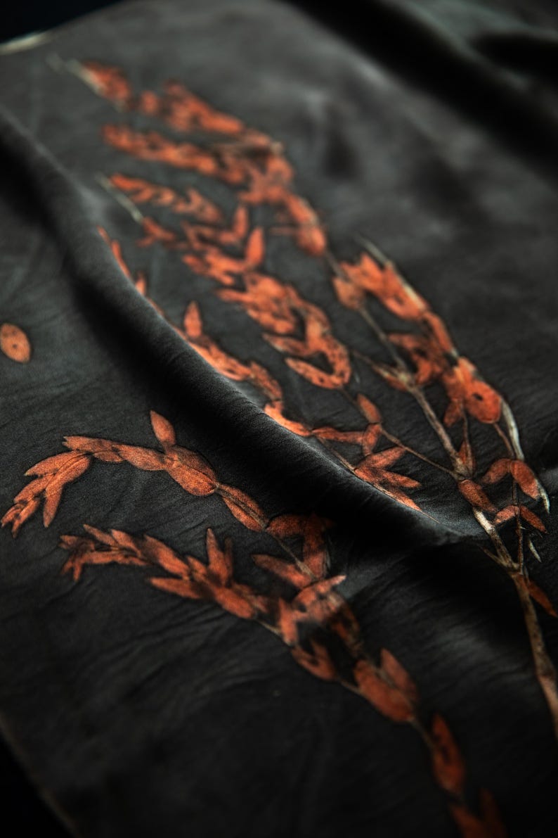 May include: A dark charcoal gray fabric featuring a botanical print of elongated, rust-colored leaves and stems. The material has a soft, slightly wrinkled texture with folds that create depth and shadows across the surface of the textile.