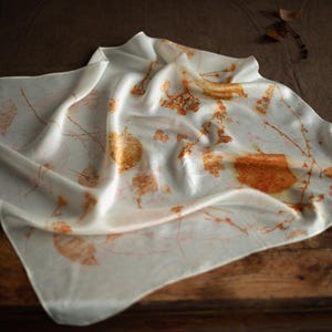 May include: A square off-white scarf featuring a botanical print with scattered orange and brown leaf and twig patterns. The fabric has a smooth, lustrous finish and is draped in soft folds, displaying a natural, organic design across the surface.