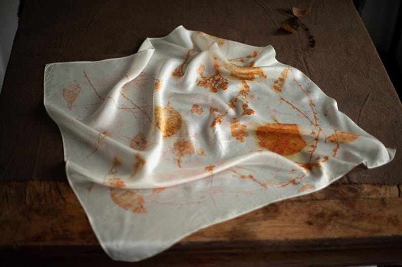 May include: A square off-white scarf featuring a botanical print with scattered orange and brown leaf and twig patterns. The fabric has a smooth, lustrous finish and is draped in soft folds, displaying a natural, organic design across the surface.