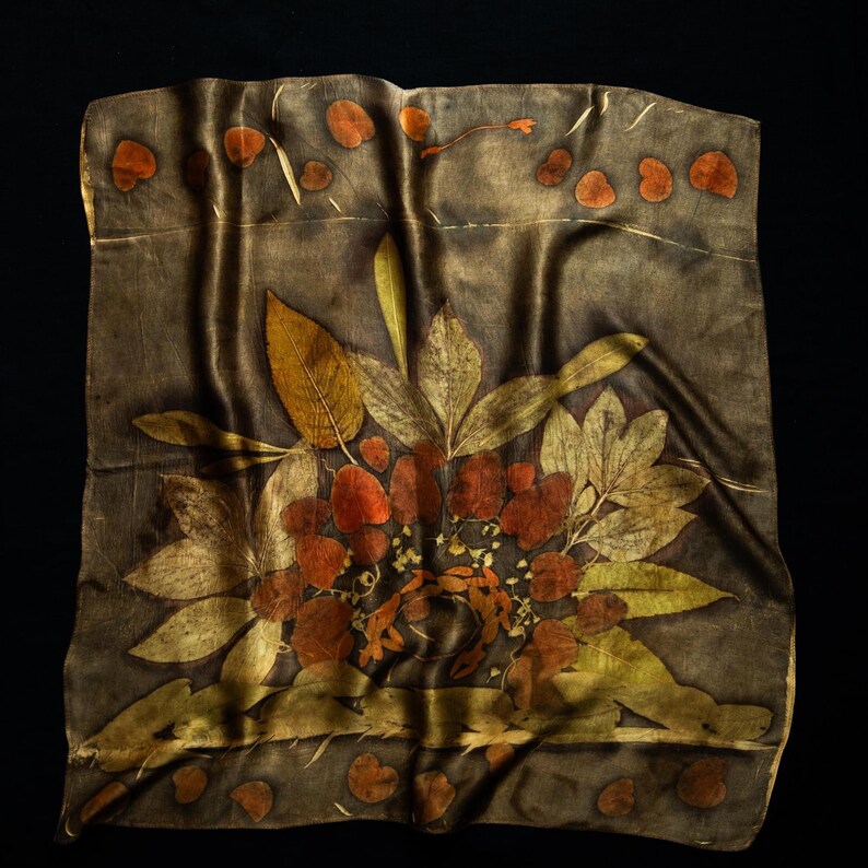 May include: A square silk scarf featuring a dark charcoal background with a botanical print of pressed leaves in shades of rust, copper, and gold. The design includes a central cluster of foliage and a border of small, scattered leaf motifs.