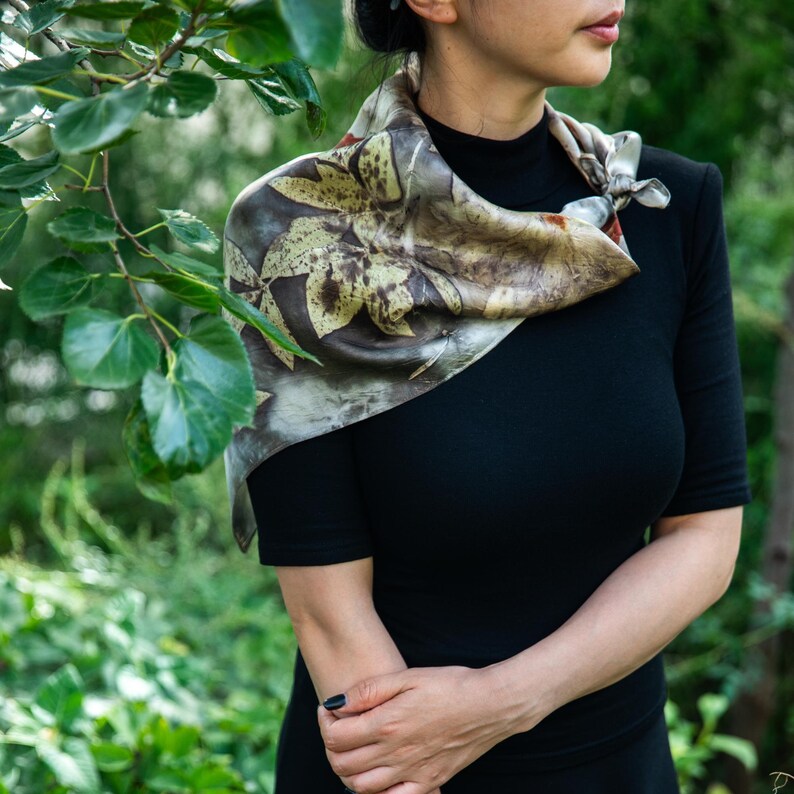 May include: A square silk scarf featuring a botanical print with dark brown and muted green leaf patterns on a light grey background. The scarf is tied in a knot at the side and is worn over a solid black short-sleeved top with a high neckline.