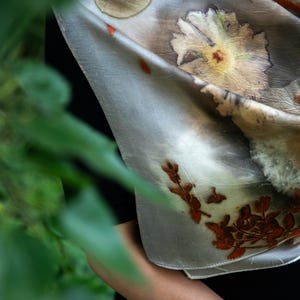 May include: A light gray silk scarf featuring botanical prints of large white flowers, green leaves, and small reddish-brown sprigs. The fabric has a smooth texture with visible natural plant imprints scattered across the surface in a soft, muted color palette.