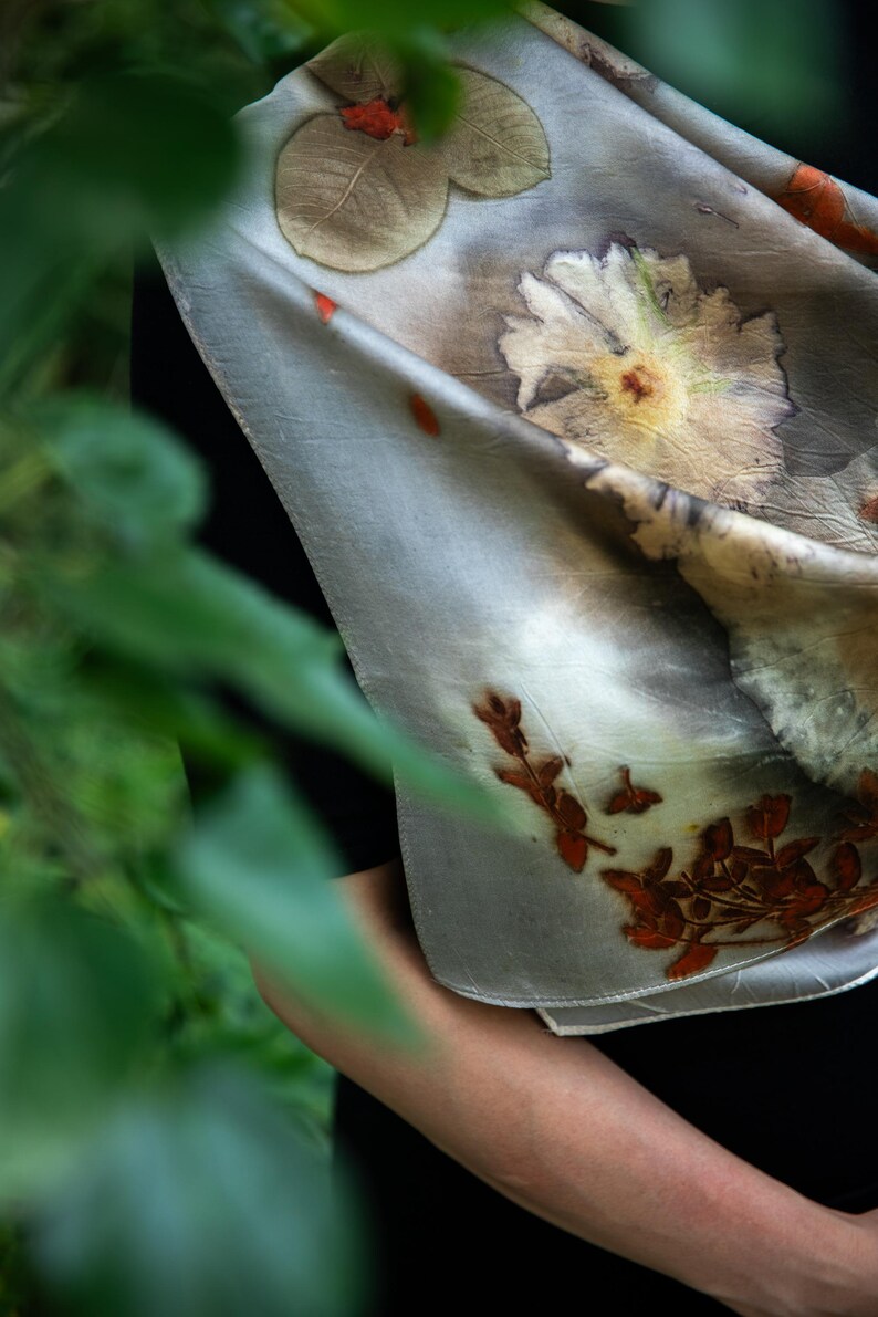 May include: A light gray silk scarf featuring botanical prints of large white flowers, green leaves, and small reddish-brown sprigs. The fabric has a smooth texture with visible natural plant imprints scattered across the surface in a soft, muted color palette.