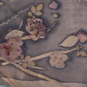May include: A rectangular piece of fabric featuring a botanical print with dark grey and muted purple tones. The design displays pressed flowers, stems, and leaves in light beige and soft pink hues, creating a natural, organic pattern across the surface.