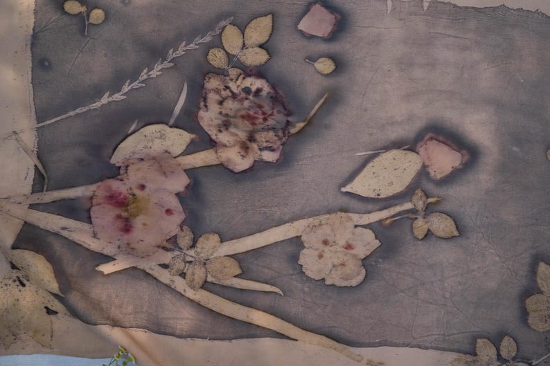 May include: A rectangular piece of fabric featuring a botanical print with dark grey and muted purple tones. The design displays pressed flowers, stems, and leaves in light beige and soft pink hues, creating a natural, organic pattern across the surface.