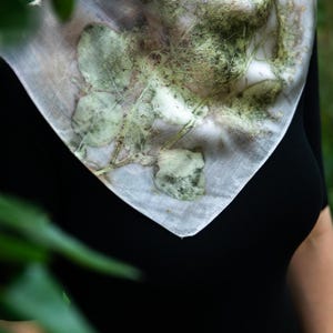 May include: A square scarf featuring a botanical print with green leaf and stem patterns on a light, off-white background. The fabric displays natural, organic shapes and textures created by plant imprints, draped around the neck area.