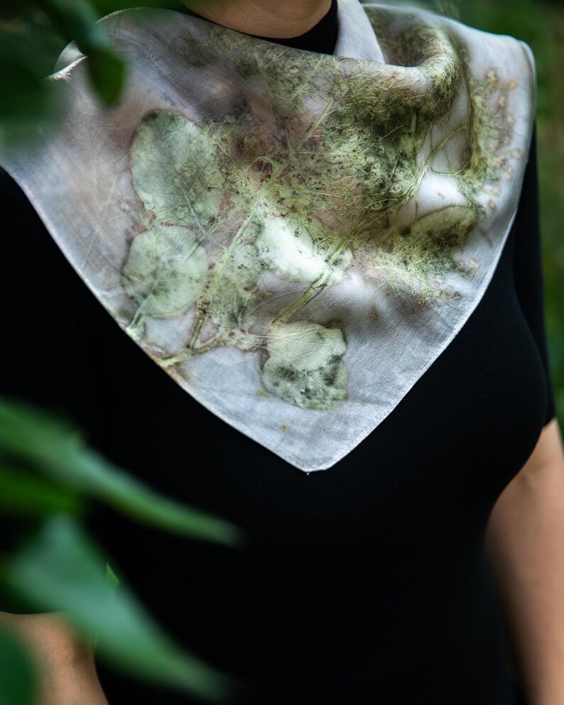 May include: A square scarf featuring a botanical print with green leaf and stem patterns on a light, off-white background. The fabric displays natural, organic shapes and textures created by plant imprints, draped around the neck area.