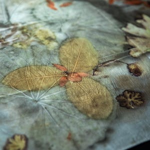 May include: A piece of fabric featuring a botanical print with pressed leaves and flowers in shades of muted green, brown, and yellow. The textile displays intricate leaf vein patterns and organic shapes transferred onto the surface.