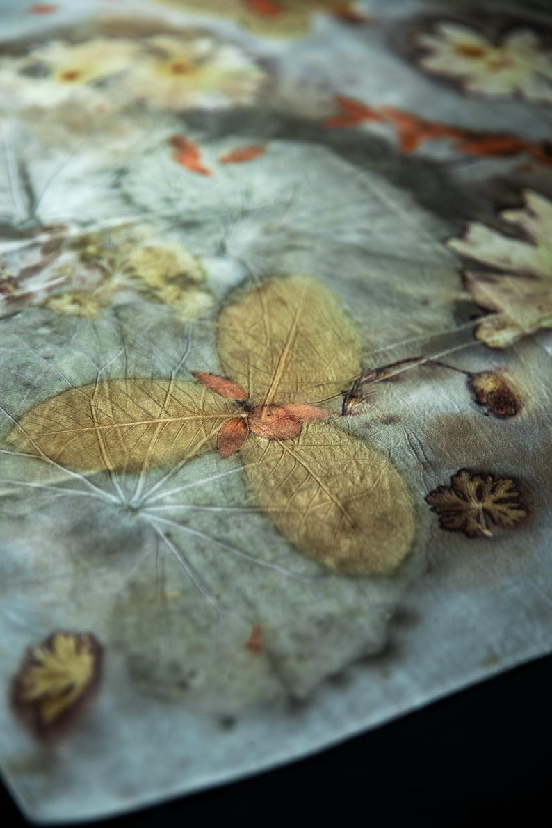 May include: A piece of fabric featuring a botanical print with pressed leaves and flowers in shades of muted green, brown, and yellow. The textile displays intricate leaf vein patterns and organic shapes transferred onto the surface.